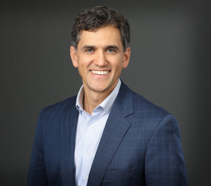Dr. Robert Thomas, a medical doctor, is smiling warmly in a blue suit and light shirt against a dark background.
