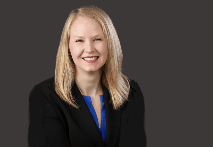 A woman with long blonde hair, wearing a black blazer and blue top, smiles against a plain gray background, embodying the professionalism often seen at Minnesota hospitals like EPPA Health.