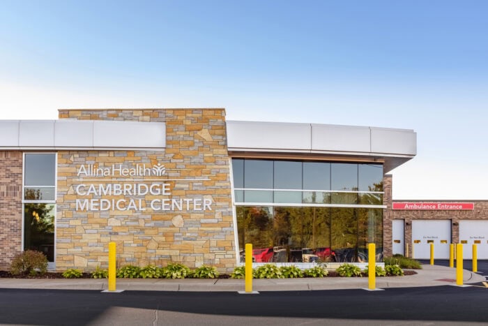 Exterior view of Allina Health Cambridge Medical Center includes the ambulance entrance on the right and stone facade with logo on the left, a proud part of Minnesota Hospitals served by expert Emergency Room Physicians.