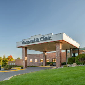 A brick hospital and clinic building under the clear blue Minnesota sky features a covered entrance, lush surrounding greenery, and top-notch emergency room physicians from EPPA Health ready to provide exceptional care.