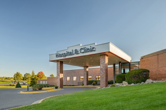A brick hospital and clinic building under the clear blue Minnesota sky features a covered entrance, lush surrounding greenery, and top-notch emergency room physicians from EPPA Health ready to provide exceptional care.