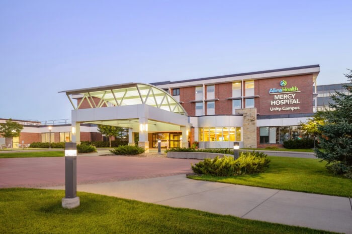 Front view of a modern hospital building with a large entrance, where "Allina Health Mercy Hospital Unity Campus" proudly stands. Surrounded by grass and pathways, this Minnesota hospital also houses EPPA Health's expert Emergency Room Physicians, ensuring exceptional care.