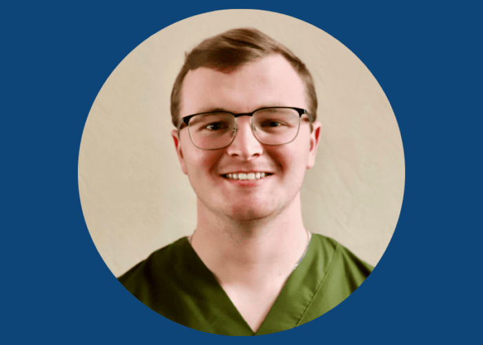 A person with short hair and glasses is smiling, wearing green scrubs against a plain beige background within a blue circular frame, representing the dedicated Emergency Room Physicians from Minnesota Hospitals.
