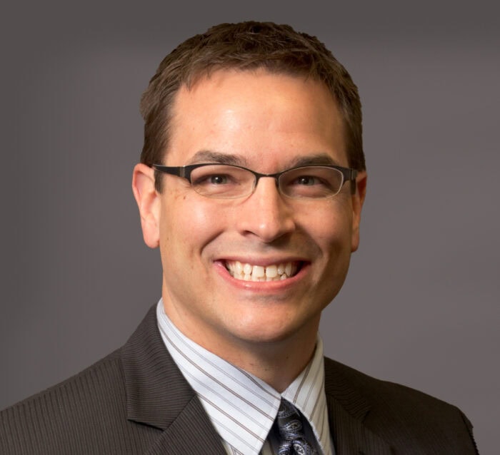 A person wearing glasses, a pinstripe suit, a striped shirt, and a patterned tie smiles at the camera against a gray background, exuding the confidence of an Emergency Room Physician from Minnesota Hospitals.