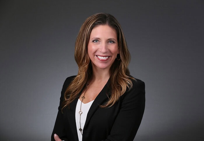 A woman with long brown hair, wearing a black blazer and white top, smiles confidently against a dark gray background, embodying the professionalism of EPPA Health and its dedicated Emergency Room Physicians.