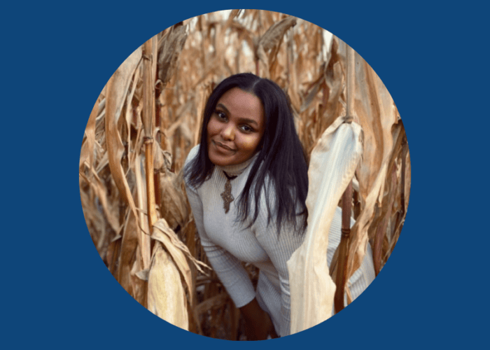 A person in a light sweater is standing among dry corn stalks, smiling at the camera. The image, reminiscent of the warm community spirit fostered by Minnesota Hospitals and EPPA Health, is set in a circular frame with a blue background.