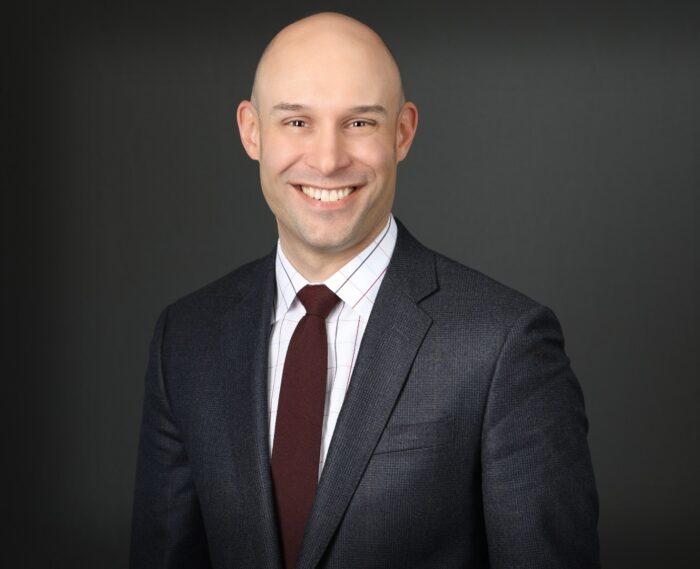 A person in a suit and tie smiling against a dark background, embodying the professionalism of Minnesota Hospitals.