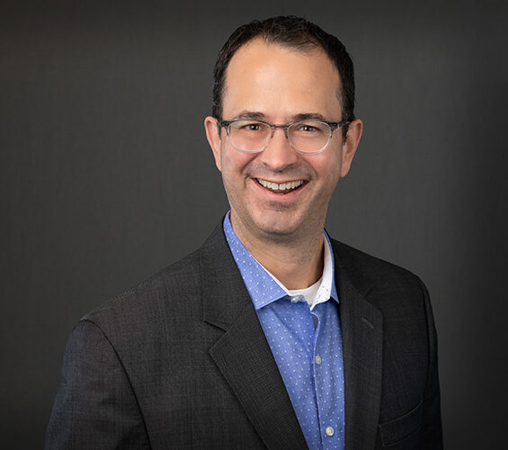 Smiling man in a suit and glasses with a blue shirt stands against a dark gray background, embodying the professionalism of Minnesota Hospitals.