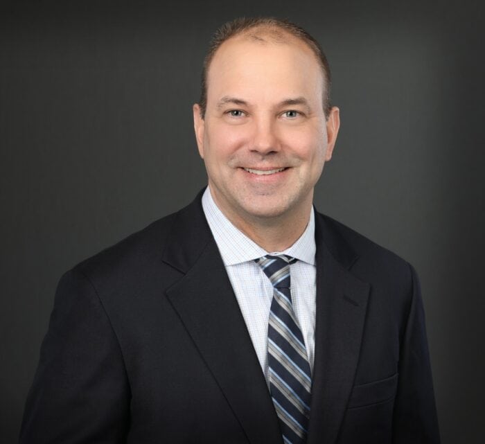 A man in a suit and tie smiles against a dark background, embodying the professionalism of EPPA Health found in Minnesota Hospitals.