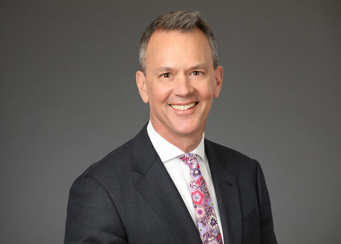 A man in a gray suit, white shirt, and floral tie smiles against a plain gray background, embodying the professionalism of Minnesota Hospitals associated with EPPA Health.