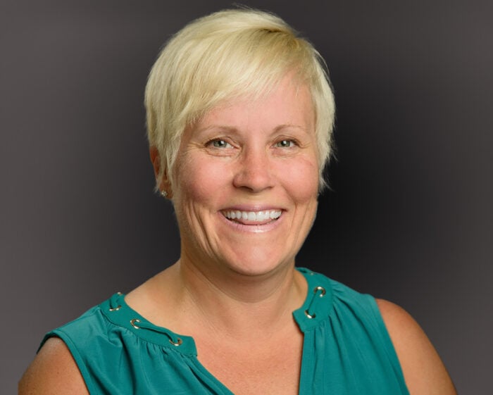A woman with short blonde hair is smiling, wearing a sleeveless teal top against a dark gray background, embodying the compassion and dedication of EPPA Health professionals.