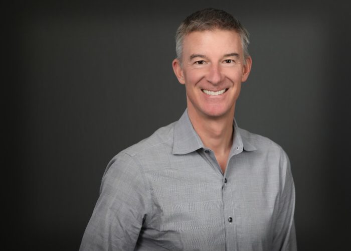 A man with short gray hair smiles while wearing a light gray button-up shirt, embodying the professionalism of an Emergency Room Physician against a plain dark background.