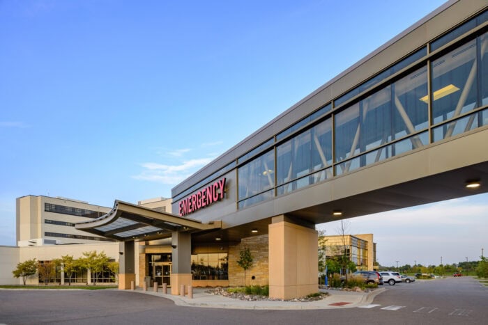 Exterior view of a Minnesota hospital entrance with a prominent "Emergency" sign and an adjoining skywalk under a clear blue sky, highlighting the dedicated Emergency Room Physicians at EPPA Health.