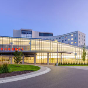 A large Minnesota Hospitals building with a glass facade and an illuminated "Emergency" sign stands against a clear evening sky, near a roundabout entrance.