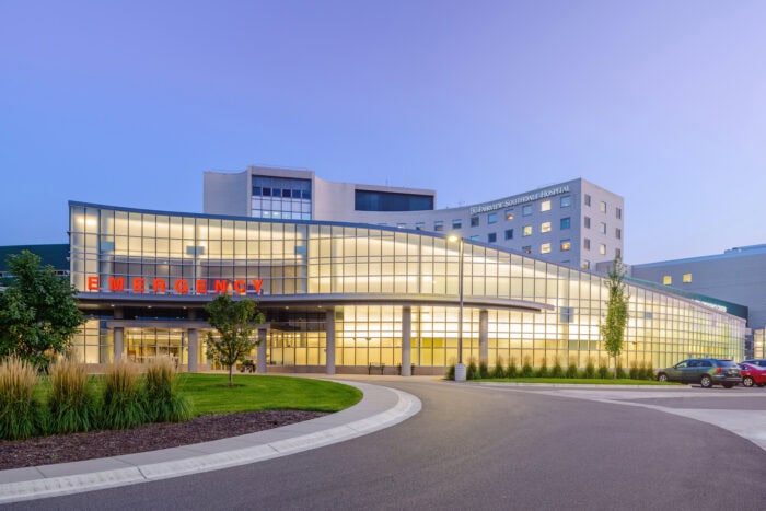 A large Minnesota Hospitals building with a glass facade and an illuminated "Emergency" sign stands against a clear evening sky, near a roundabout entrance.