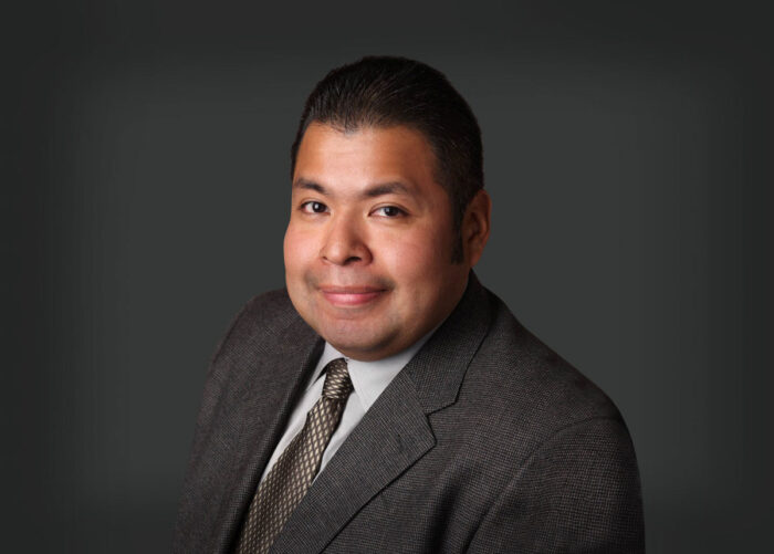 A man in a suit and tie poses against a dark gray background, smiling slightly, reminiscent of the confident demeanor often seen in Minnesota Hospitals' Emergency Room Physicians.