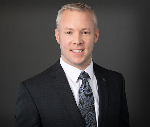 A man in a dark suit and patterned tie smiles against a gray background, embodying the professionalism seen in Minnesota hospitals.