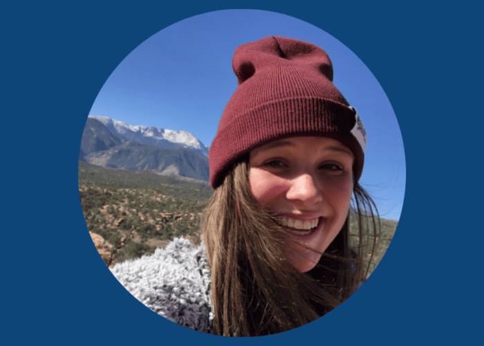 A person wearing a maroon beanie and gray sweater smiles in front of a mountainous landscape under a clear blue sky, embodying the warmth and care reminiscent of Minnesota Hospitals.