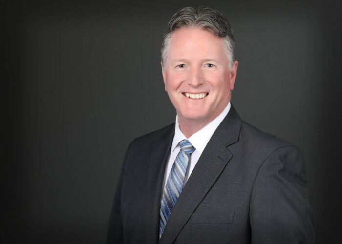 A man in a suit and tie, associated with Minnesota Hospitals, smiles against a dark gray background.