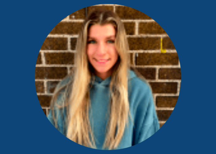 A person with long hair wearing a green hoodie stands in front of a brick wall, embodying the calmness often found among Minnesota Hospitals' Emergency Room Physicians.