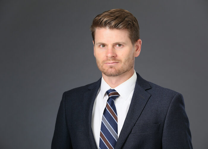 A man in a dark suit and striped tie stands confidently against a gray background, embodying the professionalism of Minnesota Hospitals and the dedication of Emergency Room Physicians.