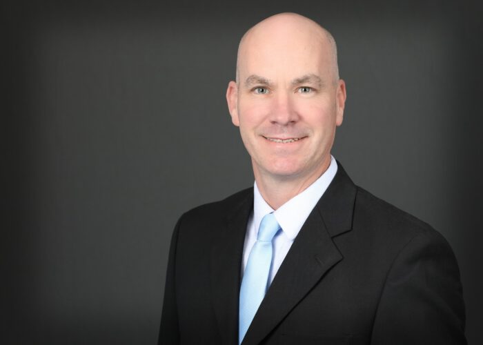 A bald man in a black suit and light blue tie, representing EPPA Health, smiles confidently against a dark gray background.