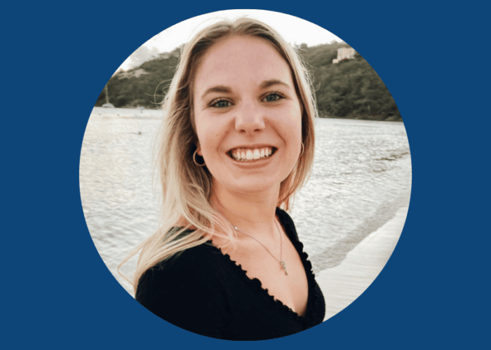 A person with long blonde hair smiles at the camera, standing by the water with a sandy shore in the background, enjoying a peaceful break from hectic days at Minnesota hospitals.