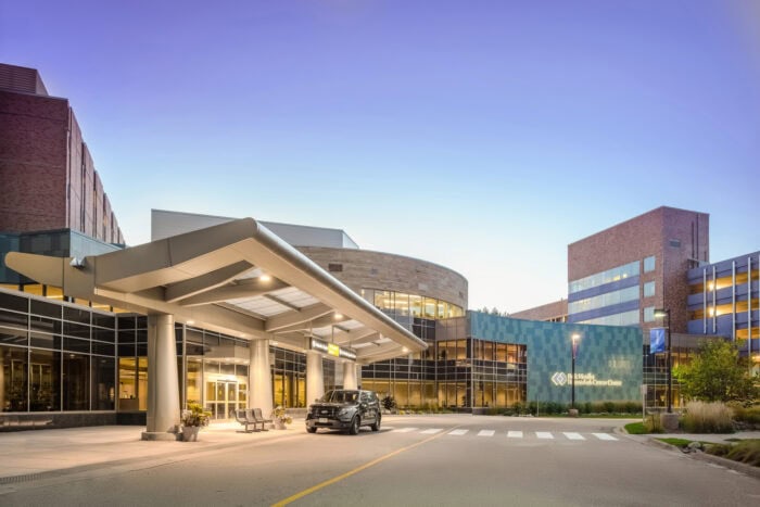 The modern hospital entrance, bearing EPPA Health signage, features a covered driveway and sleek glass facade. A parked vehicle stands nearby, while benches and greenery enhance the inviting atmosphere.