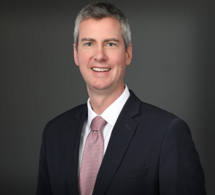 A man in a suit and tie, representing EPPA Health, smiling confidently in front of a gray background, embodying the professionalism found in Minnesota hospitals.