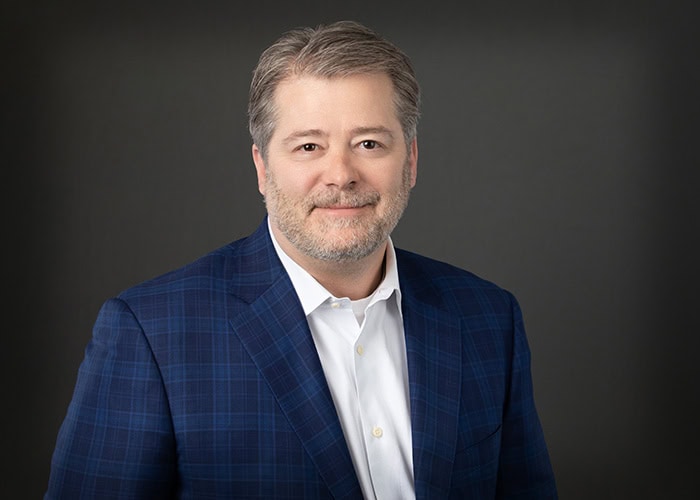 A man with gray hair and a beard, wearing a blue checked blazer and white shirt, stands confidently against a plain dark background, embodying the professionalism of Minnesota Hospitals' top Emergency Room Physicians.