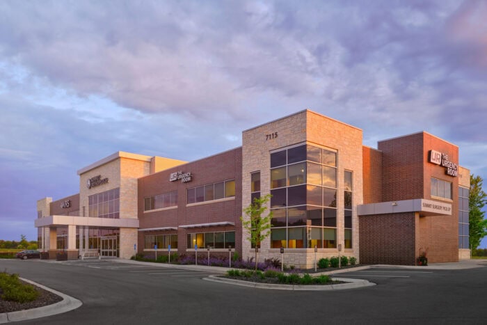 The EPPA Health facility is a modern two-story brick and stone building with large windows, surrounded by a parking lot and lush greenery. Under the cloudy Minnesota sky, its innovative design serves as a welcoming landmark for Emergency Room Physicians and visitors alike.